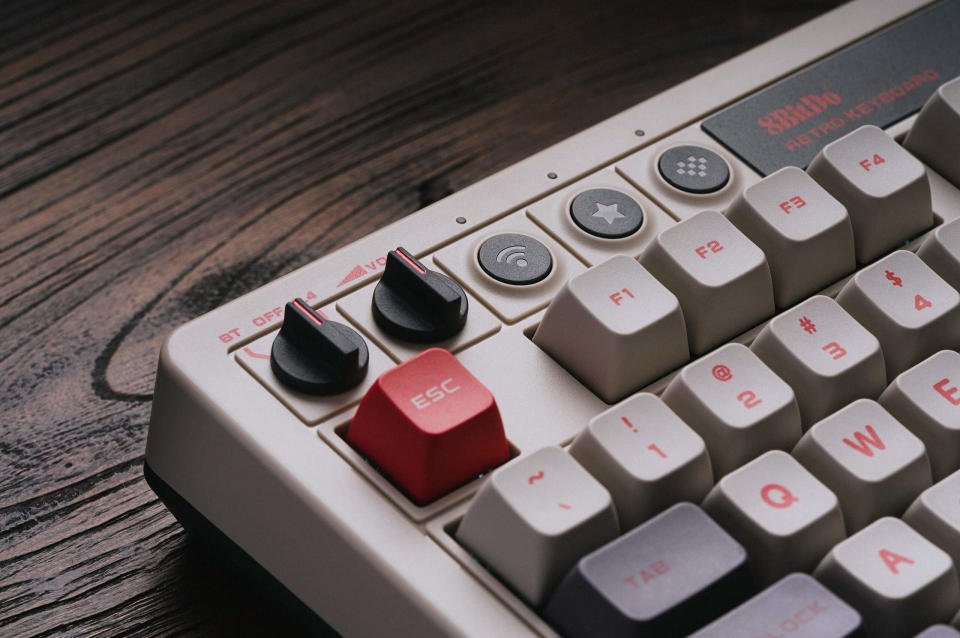 Foto produk Keyboard Mekanik Retro 8BitDo.  Tampilan jarak dekat dari kiri atas keyboard.  Ini memiliki tombol hitam dan tombol putih pudar, merah atau abu-abu tua.