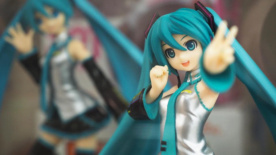 Tampilan jarak dekat dari figur Hatsune Miku.  Dia melambaikan tanda perdamaian ke arah kamera saat sosok kedua dengan pose berbeda duduk (kabur) di belakang.