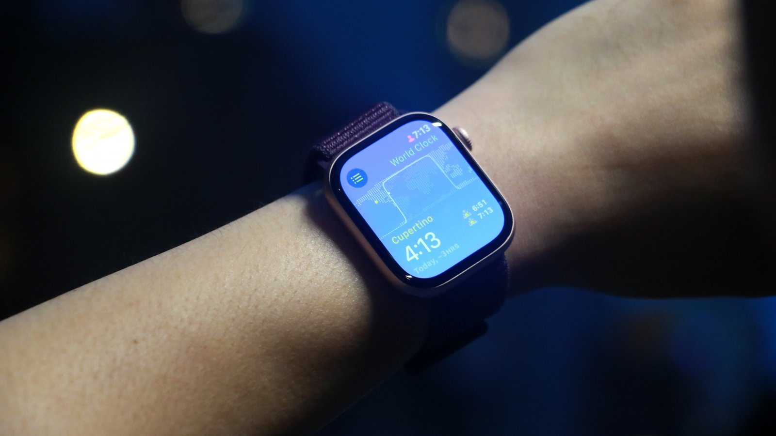 Seseorang memakai Apple Watch Series 10 di pergelangan tangan mereka. Ini menunjukkan layar biru dengan waktu dan widget lainnya. 